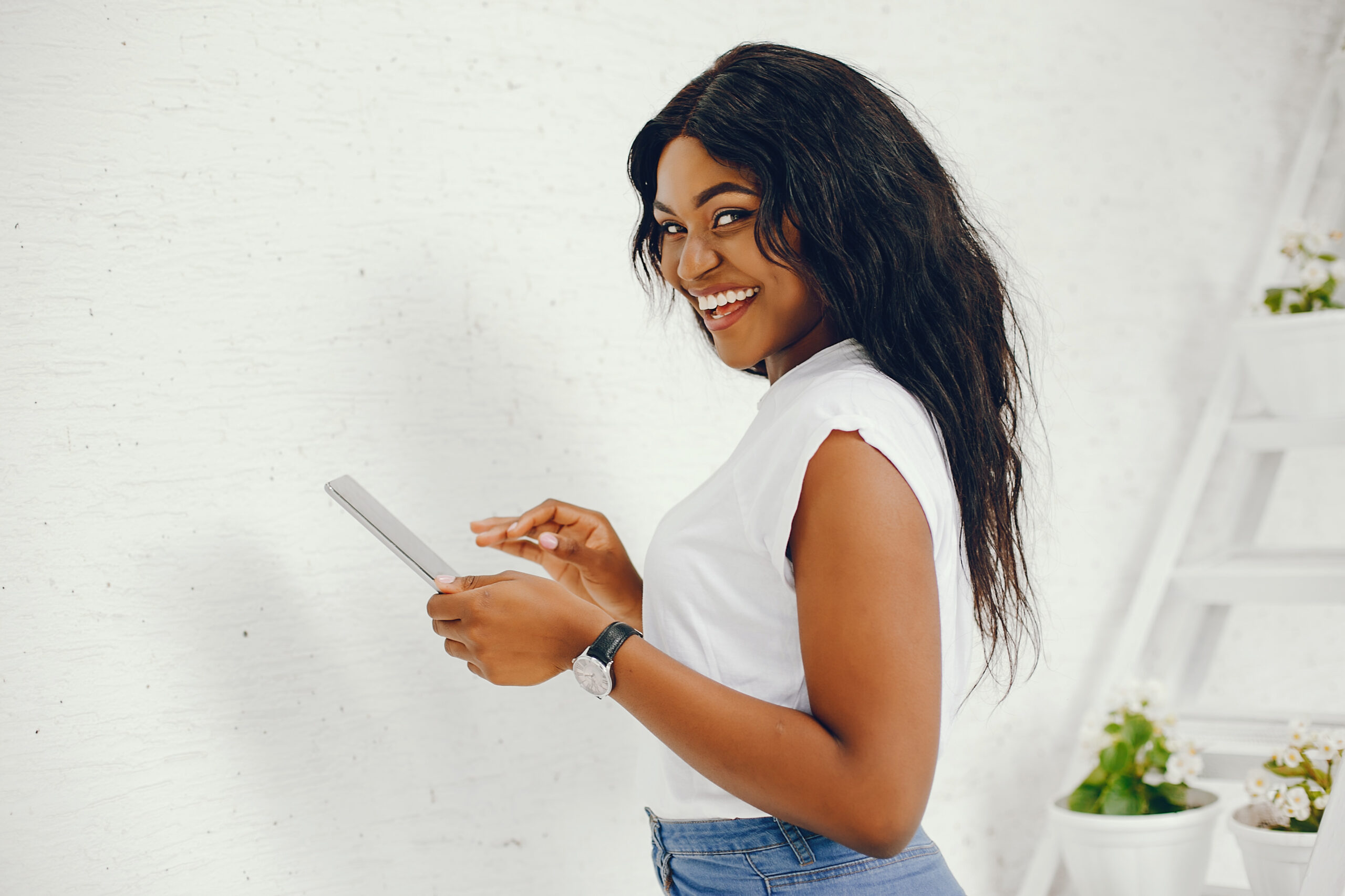 Cute black girl near white wall. Lady in a white t-shirt and blue jeans. Woman with tablet
